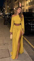 Model wearing a mustard yellow one-shoulder crop top and high slit maxi skirt set on a city street at night